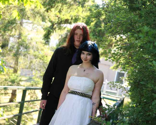 A man in a black outfit stands beside a woman in a white dress on a sunlit path surrounded by greenery.