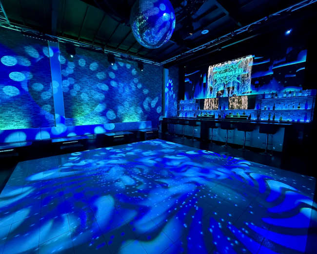 The image shows a dimly lit bar area with a shimmering disco ball, illuminated by blue lights and patterns projected on the floor and walls.