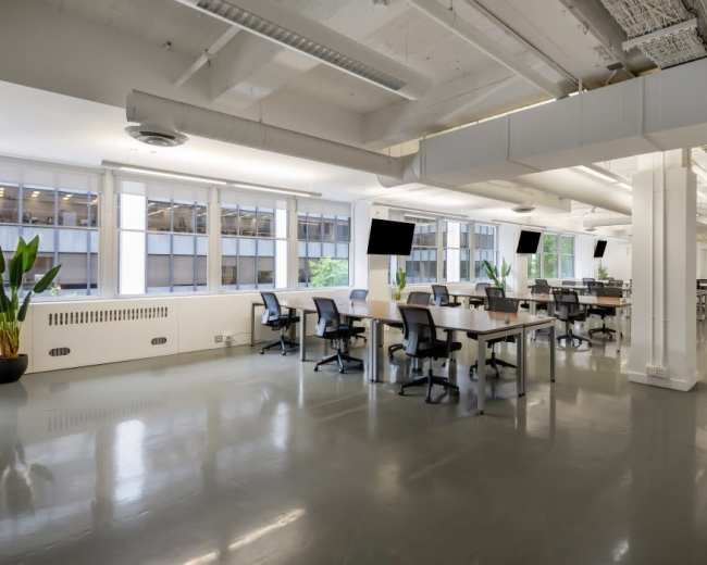 The image shows a modern office space with multiple desks, chairs, large windows, and sleek flooring.
