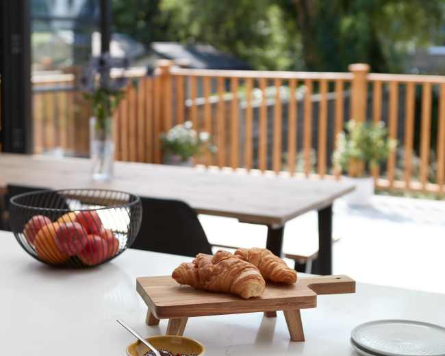 A wooden table holds a plate of croissants with a bowl of jam, while a bowl of apples sits nearby and a view of an outdoor patio is visible through large windows.