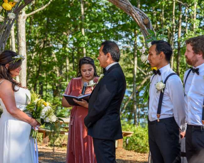 A wedding ceremony is taking place outdoors under a floral arch, with a couple exchanging vows in front of an officiant and three guests.