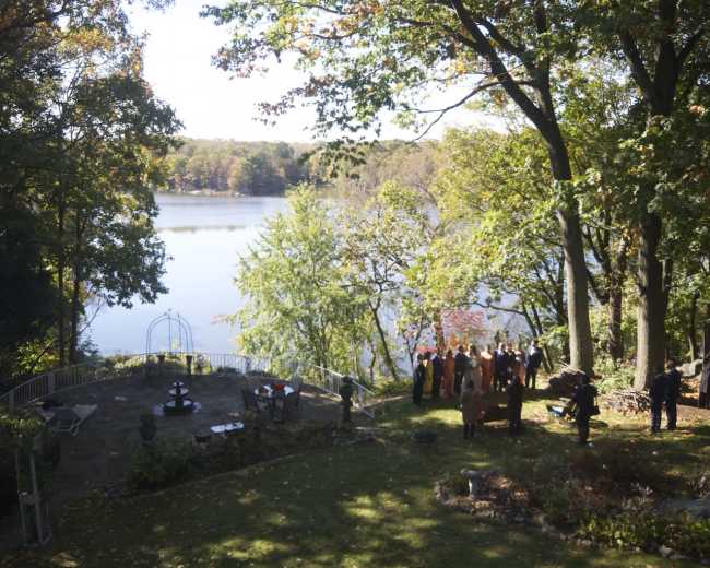 A group of people gathered by a serene lake surrounded by trees, with a landscaped yard and seating area in the foreground.