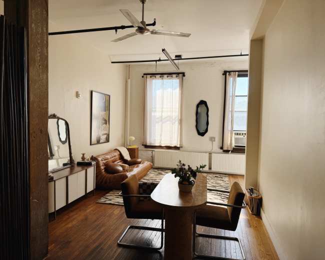 The image shows a spacious living area with a brown leather sofa, a round dining table with chairs, and large windows allowing natural light to enter.