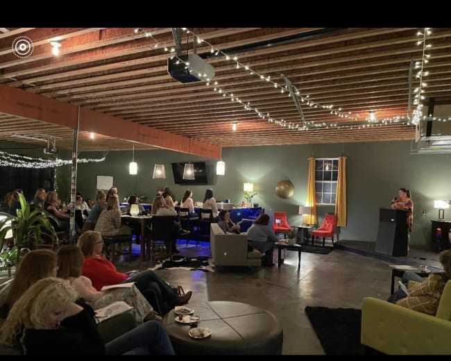A group of people are seated in a dimly lit venue with string lights, attentively facing a speaker at the front.