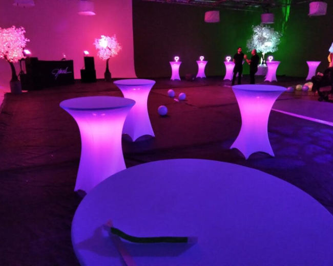 A dimly lit event space with illuminated tables, decorated trees, and a bar area in the background.