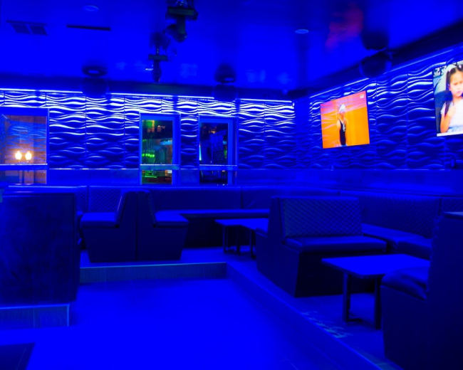 The image shows a dimly lit lounge area with blue lighting, featuring benches and tables, along with screens displaying videos on the walls.