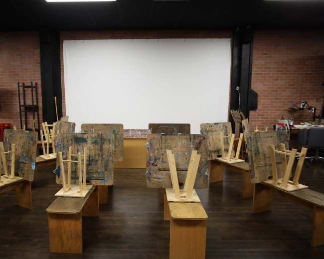 A room with several wooden easels turned upside down on wooden benches, facing a blank white screen at the front.