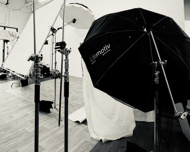 The image shows several photography equipment stands, including a black umbrella reflector, set up in a studio space with wooden flooring and white backdrop fabric.