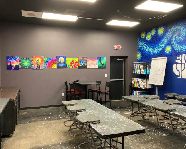 The image shows an art classroom with colorful paintings displayed on the walls, tables organized in rows, and a seating area to the side.