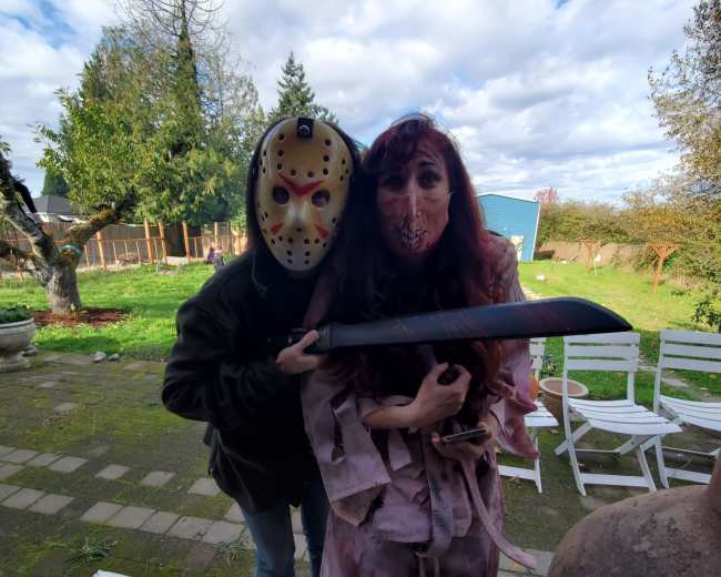 A person wearing a hockey mask stands behind a woman dressed in tattered clothing, both posing outdoors near white chairs and trees under a partly cloudy sky.