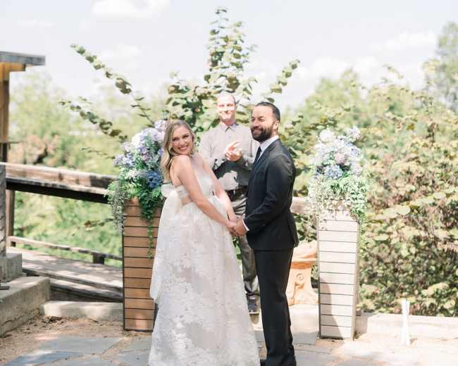 A bride and groom stand together during their wedding ceremony, with a celebrant in the background and floral arrangements on either side.
