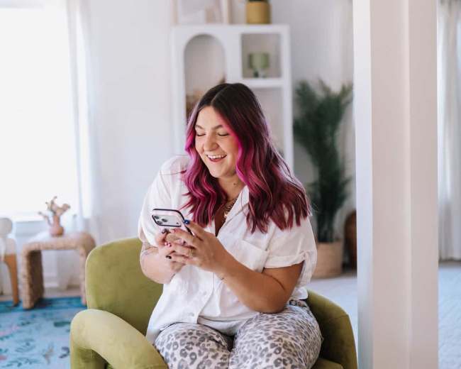 A woman with pink hair sits on a green chair, smiling while looking at her phone in a brightly lit room.