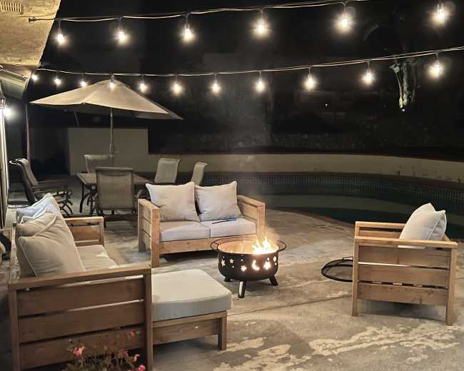 A cozy outdoor seating area features wooden chairs and a circular fire pit, illuminated by string lights above, beside a swimming pool.