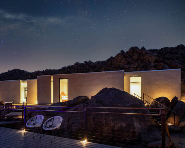 A modern building is illuminated at night, surrounded by large rocks and under a starry sky.