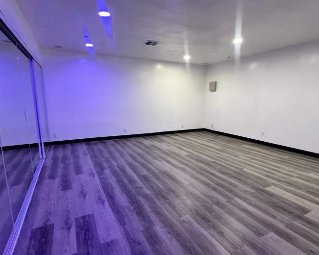 The image shows an empty room with wooden flooring and white walls, featuring recessed lighting and a large mirrored wall.