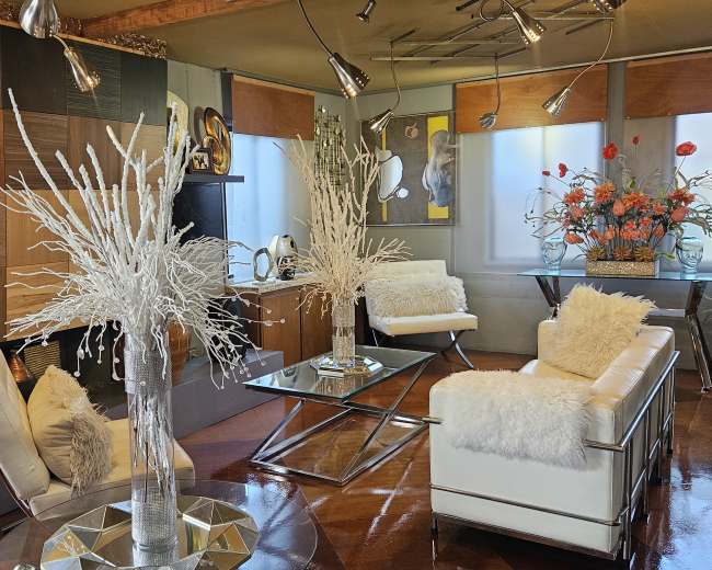 The image features a modern living room decorated with minimalist furniture, including white sofas, metallic accents, and large decorative arrangements of flowers.