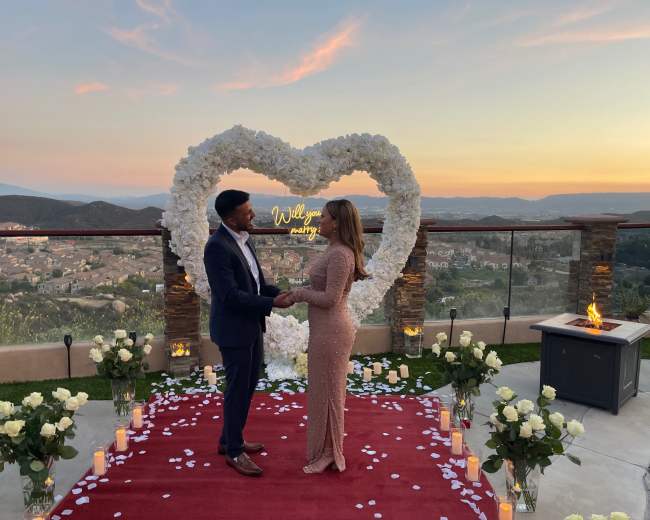 A couple stands in front of a heart-shaped floral display at sunset, surrounded by candles and rose petals on a red carpet.