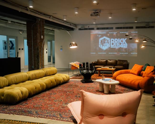 The image shows a modern lounge area featuring colorful sofas, a large area rug, a wall-mounted projector displaying "BRICK 142," and a motorcycle in the corner.