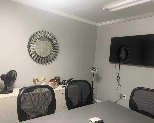 A modern office setting with a round mirror on the wall, a television, a fan, and a grey table surrounded by black mesh chairs.