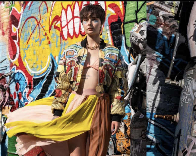 A model poses in front of a colorful graffiti wall, wearing a vibrant outfit that combines a printed jacket, a cropped top, and a multi-colored skirt.