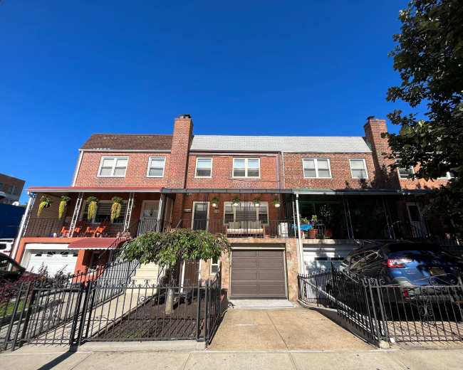A brick duplex house with a front porch, featuring a garage and a parked car, is set against a clear blue sky.