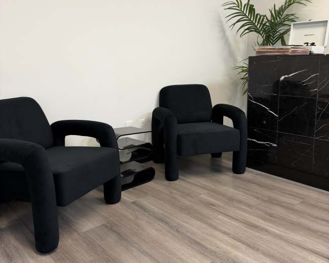 The image shows two black upholstered chairs facing each other in a room with light wooden flooring and a marble reception desk.