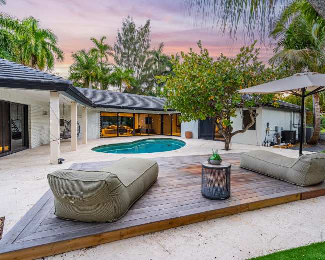 The image shows a tranquil outdoor space featuring a swimming pool, lounge seating on a wooden deck, and lush tropical landscaping.