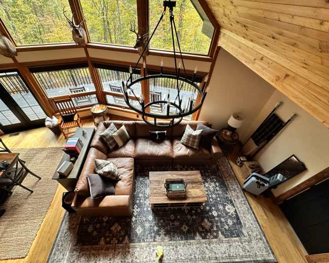 The image shows a spacious, modern living room with a large brown leather sectional sofa, a wooden coffee table, and high ceilings adorned with a circular chandelier.