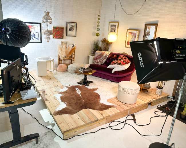 A cozy studio setup featuring a wooden platform with a cowhide rug, a burgundy sofa, and various decorative items, including a large soft light and workstations.