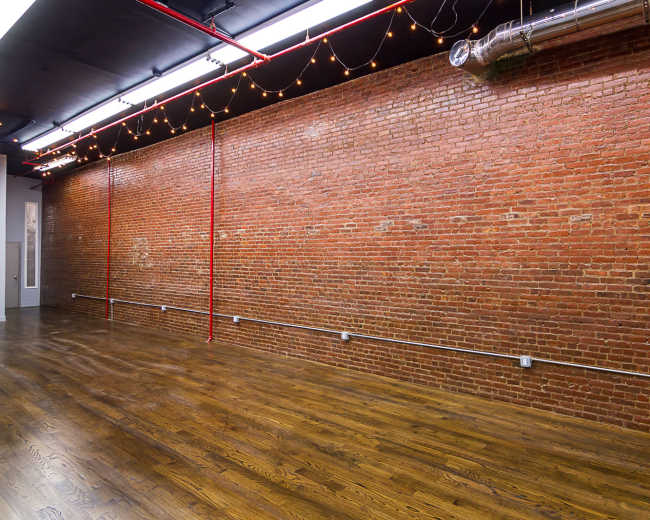 The image shows a spacious interior featuring a red brick wall and polished wooden flooring illuminated by overhead lights and string lights.
