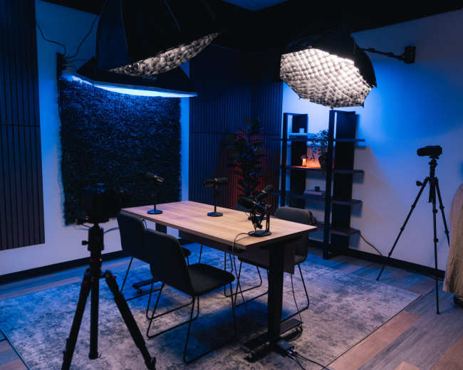 The image shows a recording studio set up with a table, chairs, microphones, and lighting equipment against a backdrop of dark textured walls.
