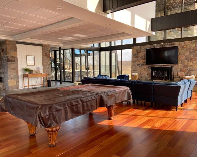 A modern indoor space featuring a pool table covered with a brown cloth, surrounded by large windows, a stone fireplace, and a seating area with blue sofas.