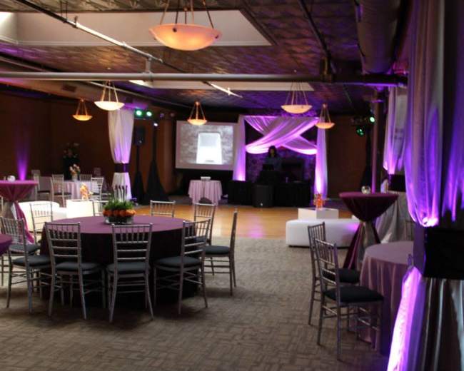 The image shows a decorated event space featuring several round tables with chairs, ambient lighting, and a DJ setup in the background.