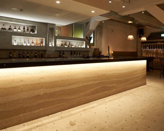 The image shows a modern bar area with a textured, light-colored counter and shelves displaying various bottles of liquor, illuminated by soft lighting.