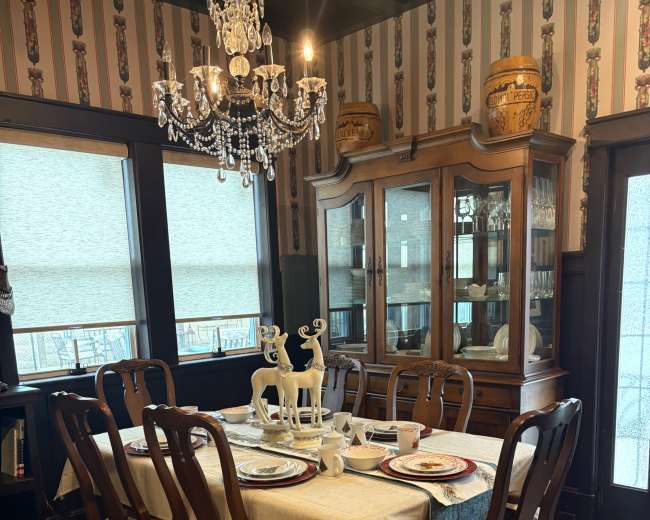 The image shows a dining room featuring a large table set with dishes, surrounded by wooden chairs, and a chandelier hanging above.