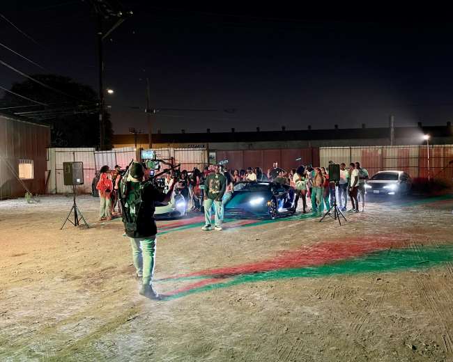 A group of people gathers around luxury cars in a lot illuminated by various lights at night.