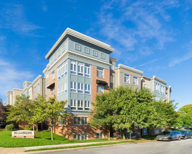 The image shows a modern, multi-story apartment building with a sign reading "20 Landbourne" in front, surrounded by trees and a clear blue sky.