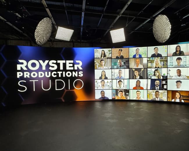 A large digital screen displays a grid of fifteen individuals in a studio setting, alongside the text "ROYSTER PRODUCTIONS STUDIO."