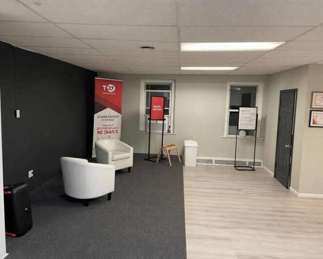 The image shows a modern office space featuring two white chairs, a promotional banner, a waste bin, and informational posters on the walls.
