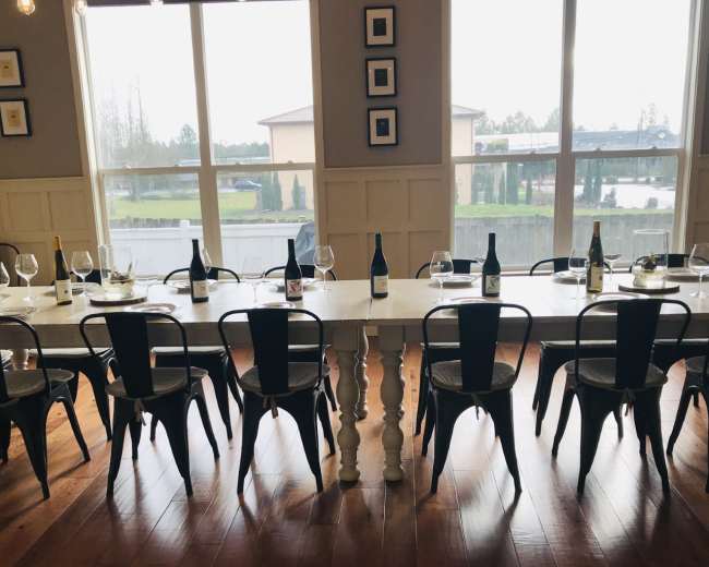 A long table set for a tasting is flanked by metal chairs, with several wine bottles and glasses arranged neatly on the table.