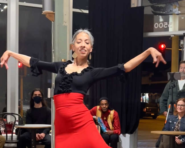 A dancer in a black top and red ruffled skirt performs in front of an audience seated at tables.