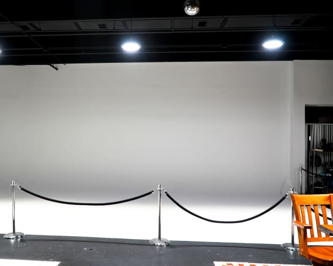 The image shows a blank studio space with a white wall, a black rope barrier, and a wooden chair in the foreground.