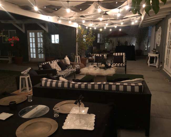 The image shows a cozy outdoor seating area with striped cushions, decorative lanterns, and string lights hanging overhead.