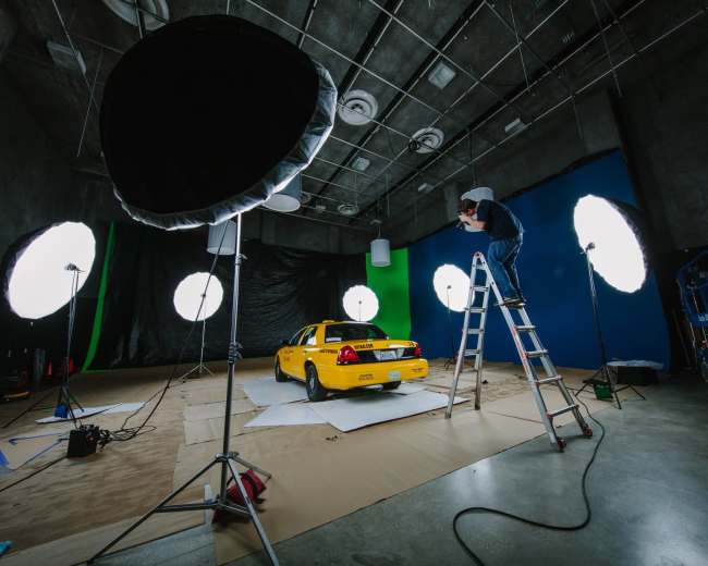 A yellow car is positioned in a studio space with large lighting equipment and a person on a ladder adjusting the lights.