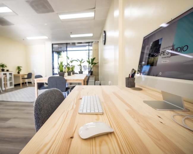 A modern office space features a wooden desk with a computer, keyboard, and mouse, surrounded by plants and furniture.