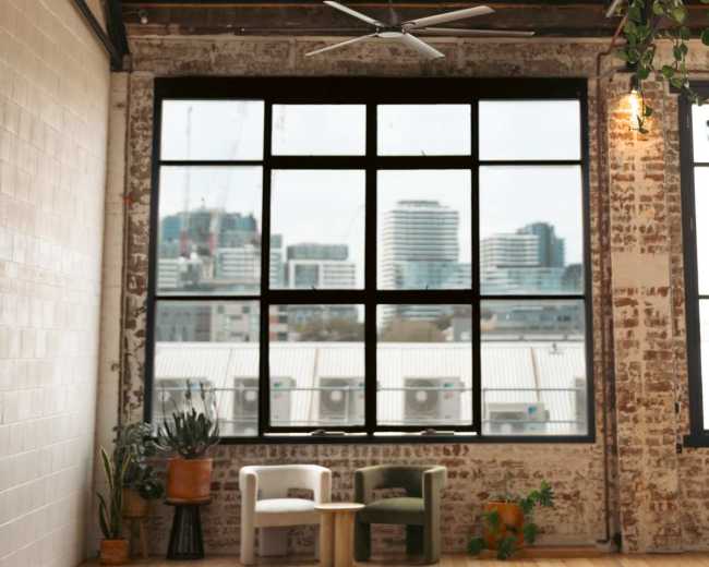 The image shows an industrial-style interior with a large, round pendant light, a ceiling fan, and a window revealing a city skyline in the background.