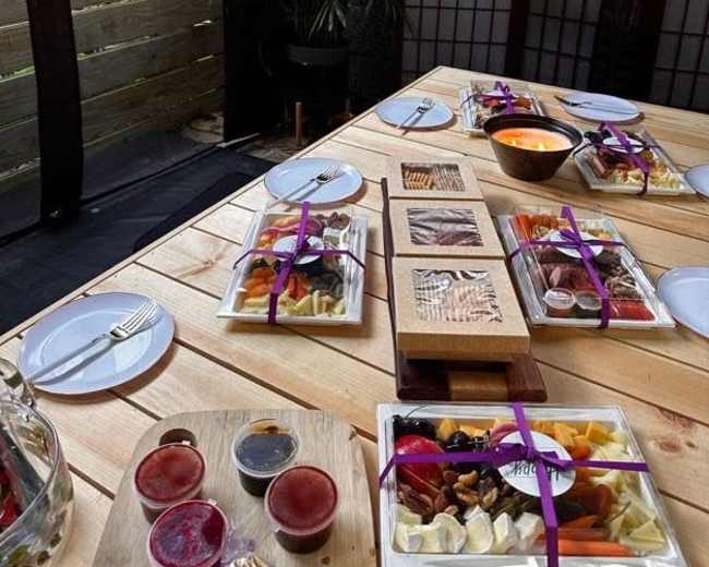 A wooden table is set outdoors with multiple plates of food, sauces, and a bowl, surrounded by partitions and greenery.