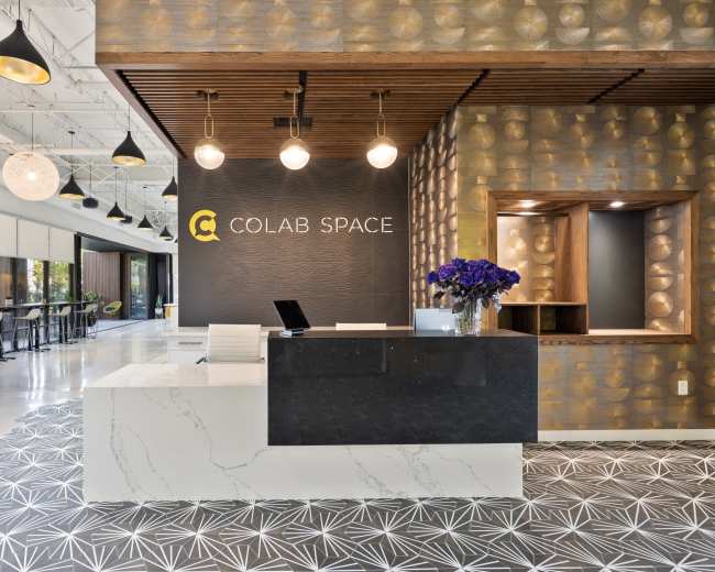 The image shows a modern reception area labeled "COLAB SPACE," featuring a sleek front desk with a dark counter, decorative wall patterns, and seating in the background.