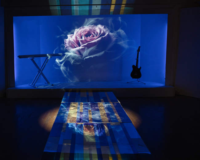 The scene features a blue-lit stage with a large projected image of a rose and a keyboard on the left, complemented by an electric guitar on the right.