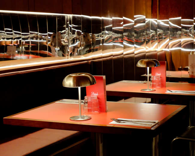 The image shows a modern restaurant interior with red tables, sleek metal lamps, and mirrored walls reflecting the surroundings.